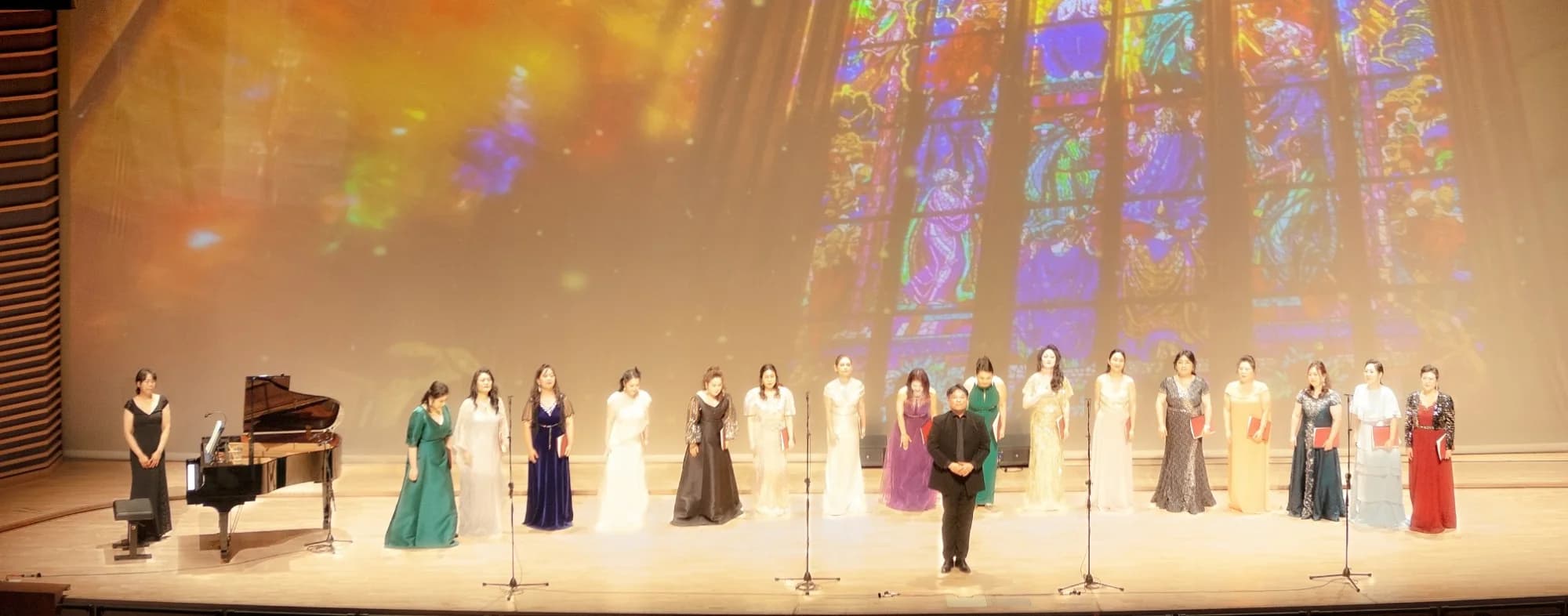 Women's vocal ensemble taking a bow at the HTUS 70th anniversary concert, under a projected stained-glass window