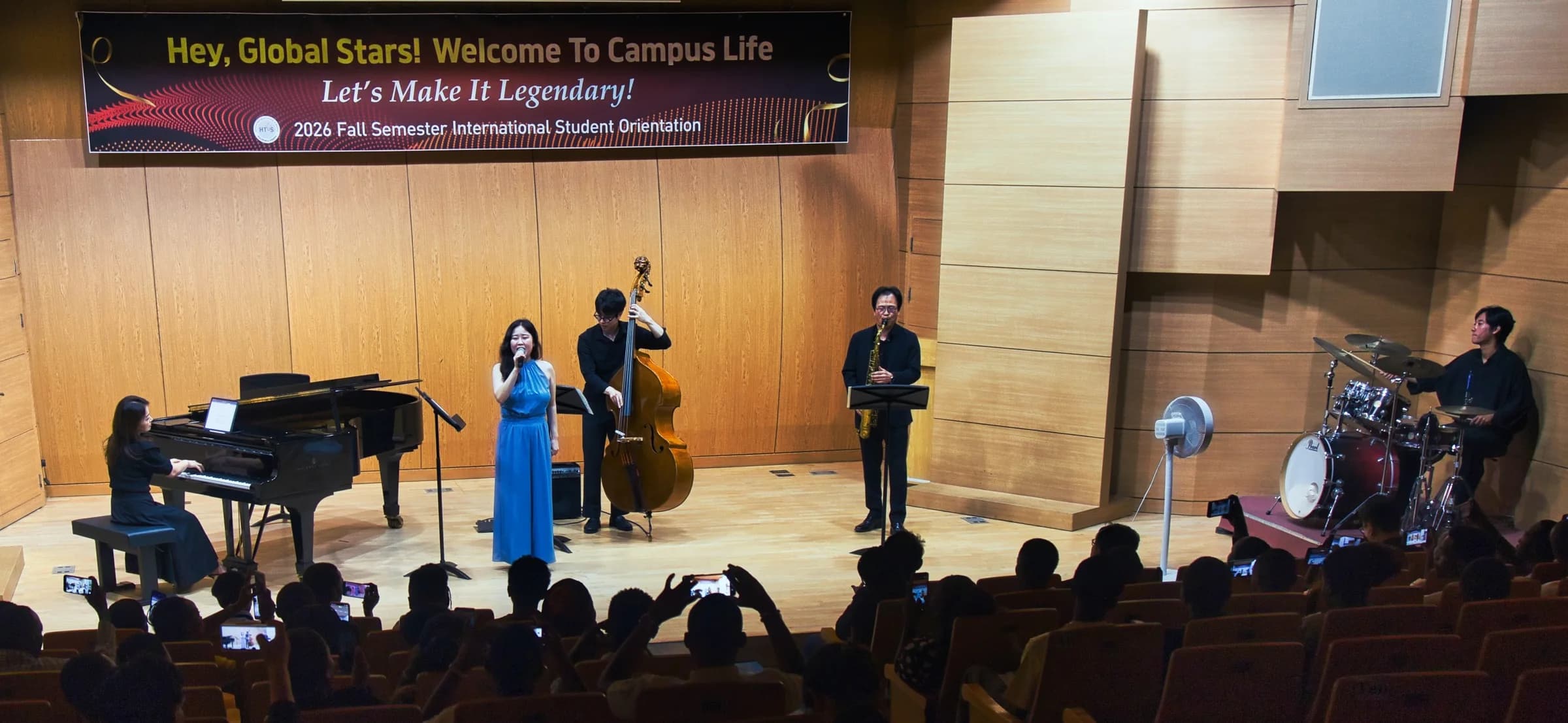 Welcome concert on stage: piano, vocalist, double bass, saxophone and drums, with students filming from their seats