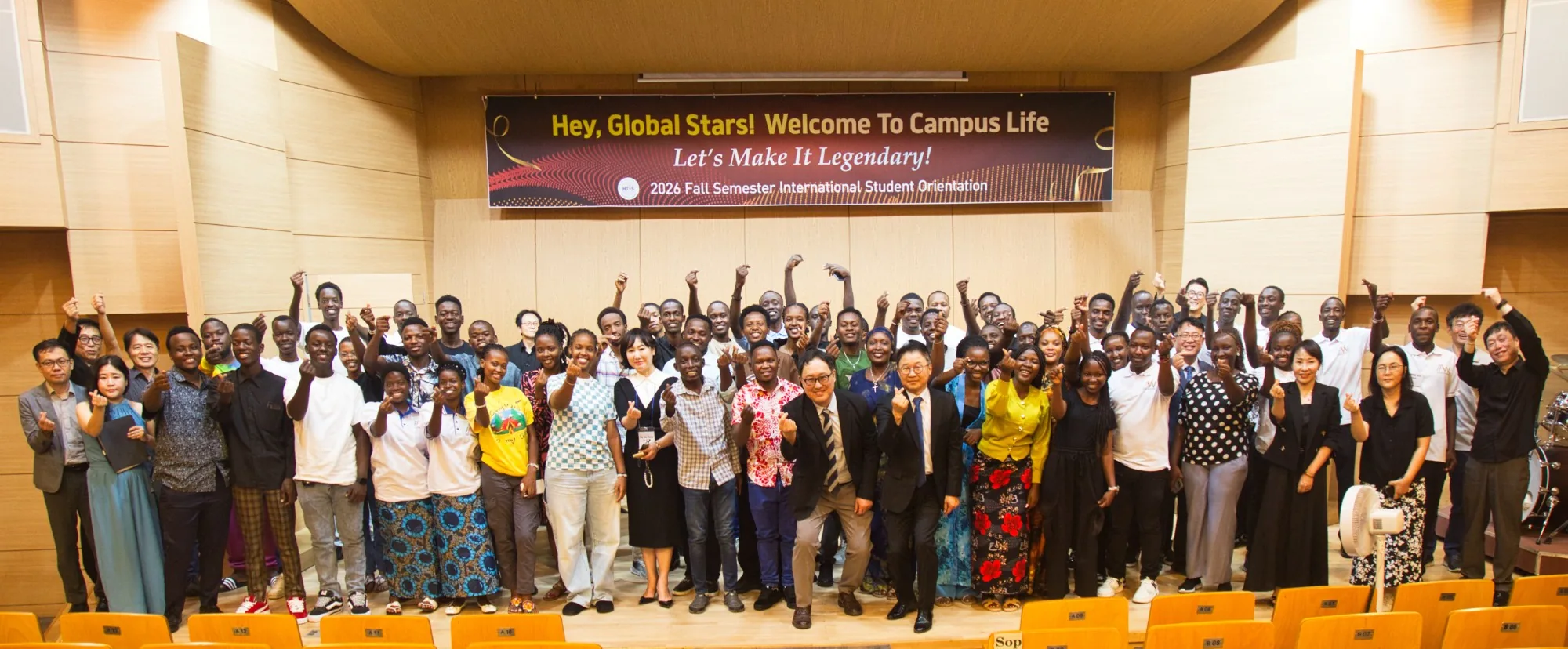 New and returning international students with faculty and staff on stage at the 2026 Fall orientation, all making the finger-heart sign